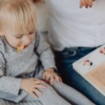 Cute toddler with ponytails enjoying a snack next to a book and parent. Cozy indoor scene.